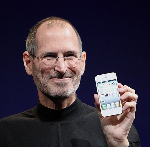 Steve Jobs smiling in a black turtleneck while holding up a white iPhone 4