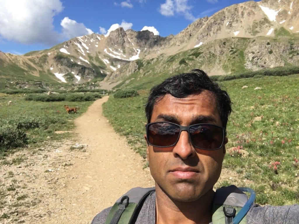 Selfie on a high alpine trail in summer, a dog trotting up the path and snow-streaked peaks behind