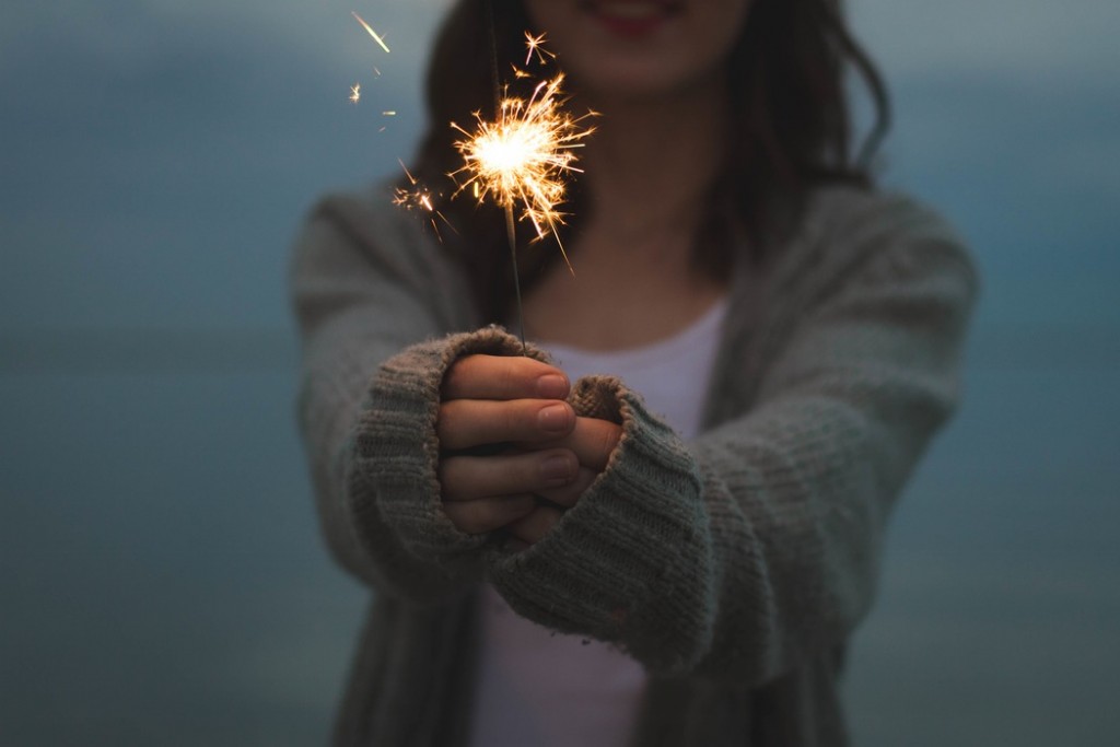 Woman in a gray cardigan holding a lit sparkler toward the camera