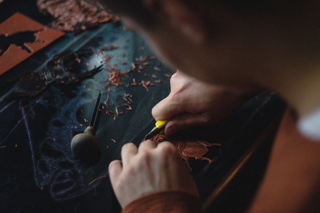 Close-up of hands carving an intricate linocut printing block, with shavings scattered across the dark surface