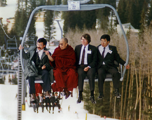 The Dalai Lama in red robes riding a ski chairlift with three suited companions above a slope