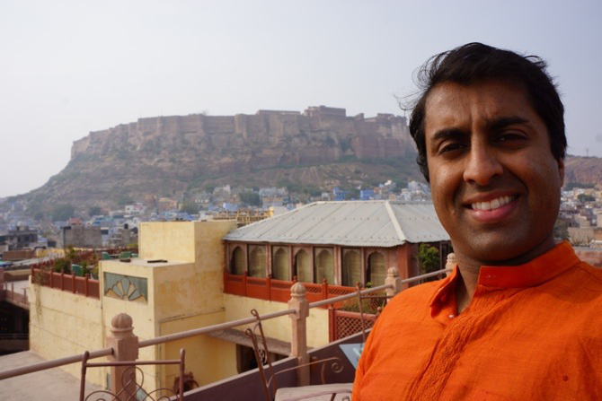 Smiling man in an orange kurta on a rooftop in Rajasthan, India, with a large hilltop fort in the distance