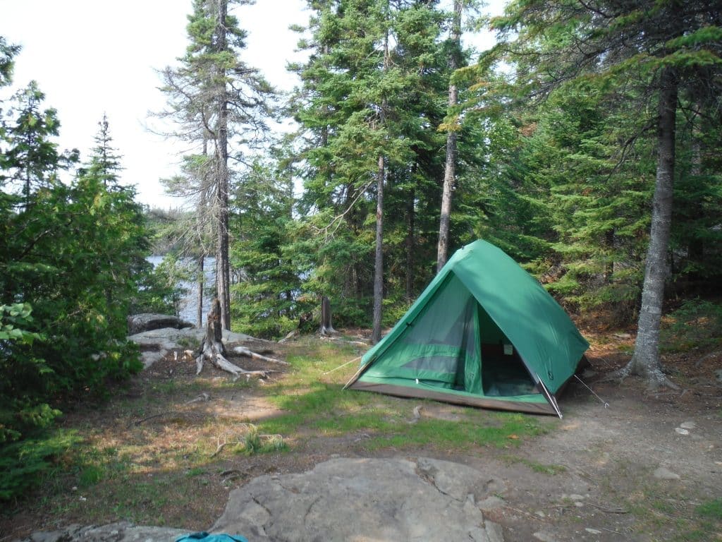 Green tent pitched on a rocky forest campsite among pines, with a lake visible through the trees