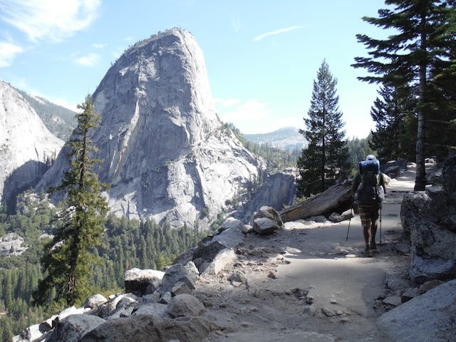 Backpacker with trekking poles on a rocky trail, a bare granite dome rising across the valley