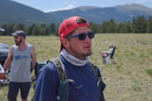 Trail runner in a backwards red cap, sunglasses, and hydration pack standing in a mountain meadow