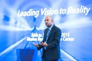 Manoj Vasudevan speaking on stage in a dark suit before a screen reading “Leading Vision to Reality”