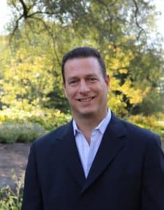 Jim Posner in a dark blazer and open-collared shirt standing outdoors among trees
