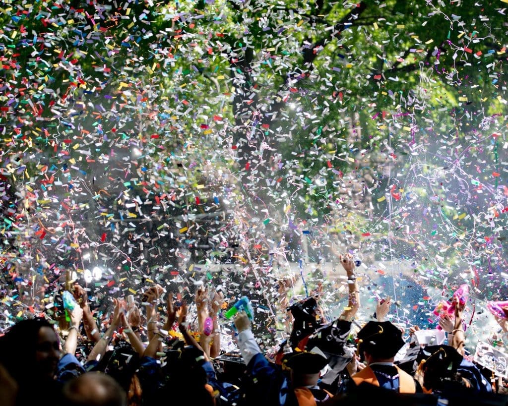 Graduates in caps and gowns raising their hands amid clouds of colorful confetti